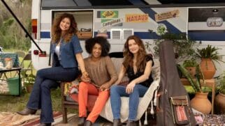Três mulheres sentadas em frente a motorhome decorado durante viagem, com plantas e violão ao lado.
