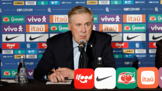 A imagem mostra o técnico Carlo Ancelotti sentado atrás de uma mesa durante uma entrevista coletiva oficial, com um painel de patrocinadores ao fundo. Carlo Ancelotti O treinador está centralizado na imagem, posicionado do peito para cima. Ele tem pele clara, cabelos grisalhos curtos penteados para o lado e uma expressão facial séria e compenetrada, olhando ligeiramente para a esquerda. Ele veste um terno azul-marinho, camisa social azul-clara e gravata escura. Suas mãos estão sobre a mesa, segurando uma caneta preta. À sua frente, há um microfone pedestal direcionado para sua boca. Elementos sobre a Mesa Na mesa de cobertura escura, em primeiro plano à esquerda, há uma garrafa de vidro transparente com água e um copo de vidro virado de cabeça para baixo sobre um guardanapo. Na borda inferior da imagem, quatro placas quadradas com cantos arredondados exibem logotipos de patrocinadores em destaque: iFood (vermelha), Nike (branca), Guaraná Antarctica (verde) e Itaú (laranja). O Cenário ao Fundo O plano de fundo é composto por um painel publicitário azul repleto de logotipos repetidos de marcas patrocinadoras da Seleção Brasileira, organizados em fileiras horizontais. Entre as marcas visíveis estão Vivo, Itaú, Guaraná Antarctica, Nike, Cimed e iFood, além da inscrição do site oficial "CBF.COM.BR".