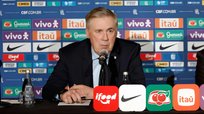 A imagem mostra o técnico Carlo Ancelotti sentado atrás de uma mesa durante uma entrevista coletiva oficial, com um painel de patrocinadores ao fundo. Carlo Ancelotti O treinador está centralizado na imagem, posicionado do peito para cima. Ele tem pele clara, cabelos grisalhos curtos penteados para o lado e uma expressão facial séria e compenetrada, olhando ligeiramente para a esquerda. Ele veste um terno azul-marinho, camisa social azul-clara e gravata escura. Suas mãos estão sobre a mesa, segurando uma caneta preta. À sua frente, há um microfone pedestal direcionado para sua boca. Elementos sobre a Mesa Na mesa de cobertura escura, em primeiro plano à esquerda, há uma garrafa de vidro transparente com água e um copo de vidro virado de cabeça para baixo sobre um guardanapo. Na borda inferior da imagem, quatro placas quadradas com cantos arredondados exibem logotipos de patrocinadores em destaque: iFood (vermelha), Nike (branca), Guaraná Antarctica (verde) e Itaú (laranja). O Cenário ao Fundo O plano de fundo é composto por um painel publicitário azul repleto de logotipos repetidos de marcas patrocinadoras da Seleção Brasileira, organizados em fileiras horizontais. Entre as marcas visíveis estão Vivo, Itaú, Guaraná Antarctica, Nike, Cimed e iFood, além da inscrição do site oficial "CBF.COM.BR".