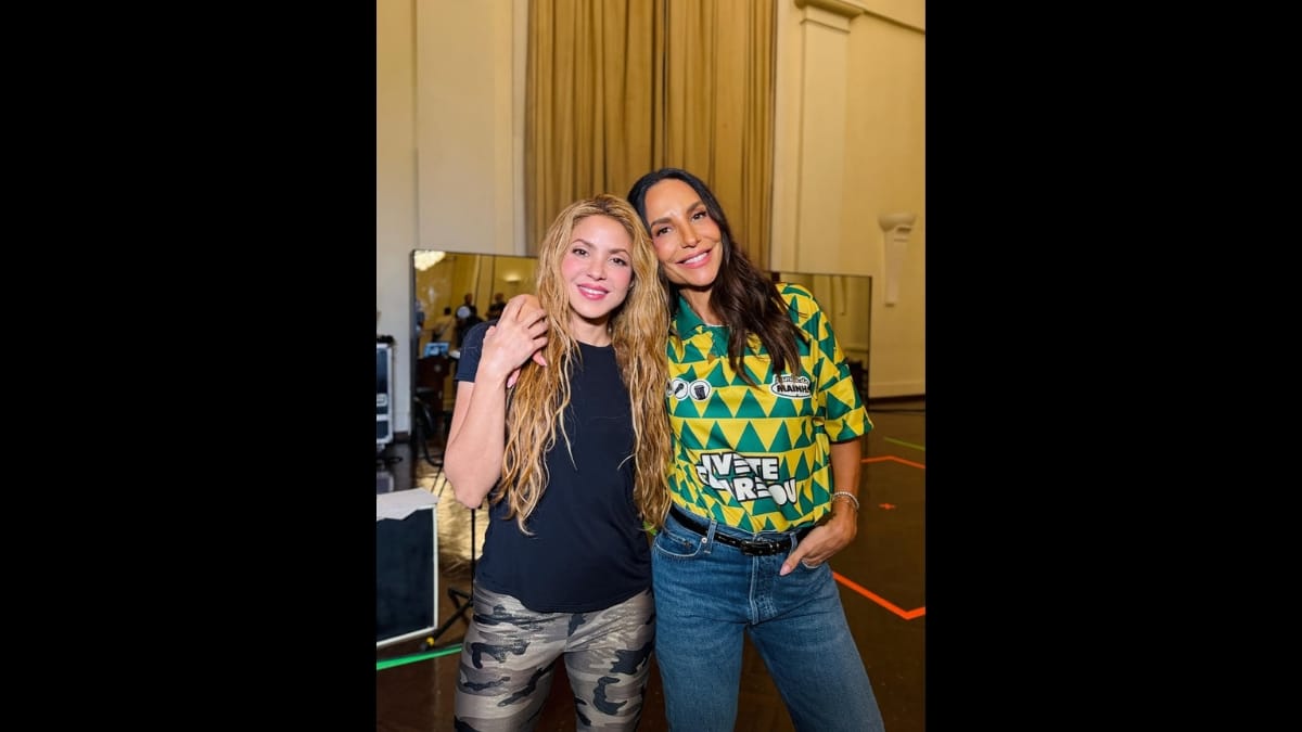 Foto colorida de Shakira e Ivete Sangalo sorrindo juntas para a câmera em um ambiente de bastidor, com Ivete Sangalo vestindo uma camisa de futebol amarela, verde e azul.
