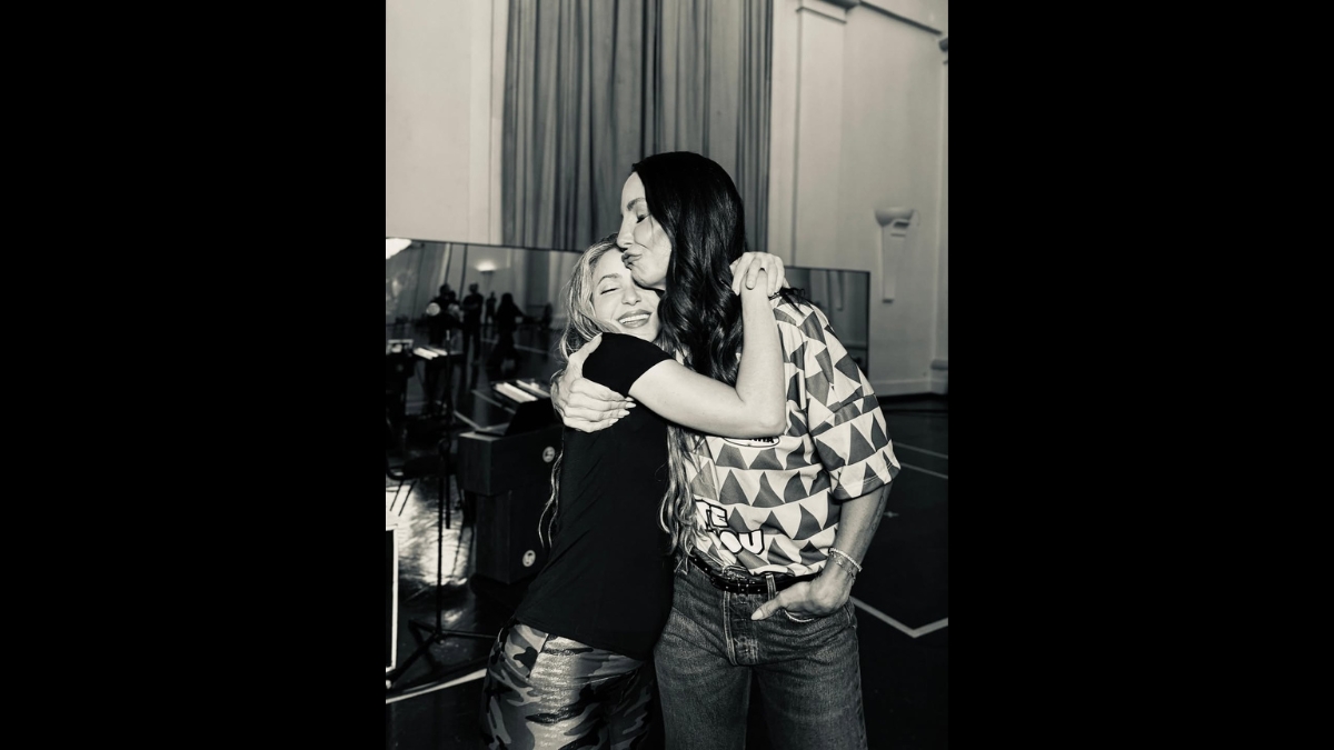 Foto em preto e branco de Shakira e Ivete Sangalo se abraçando em um estúdio ou bastidor, com Ivete Sangalo vestindo uma camisa de futebol.