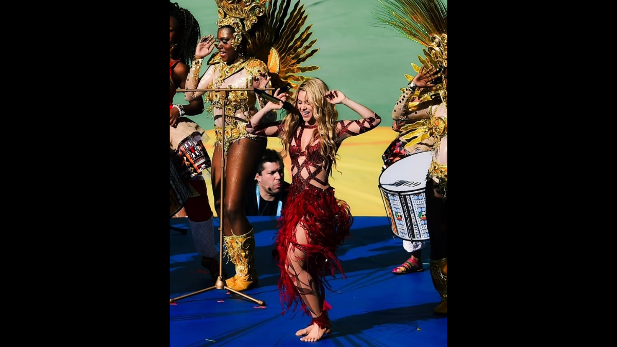 Shakira se apresentando energeticamente no palco com uma fantasia vermelha recortada e saia de franjas, cercada por dançarinos e percussionistas em fantasias douradas.