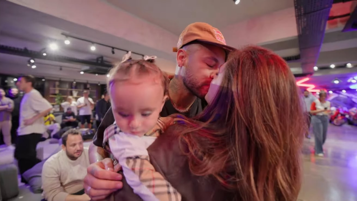 Neymar Jr. beijando o rosto de uma mulher que está segurando uma bebê no colo.