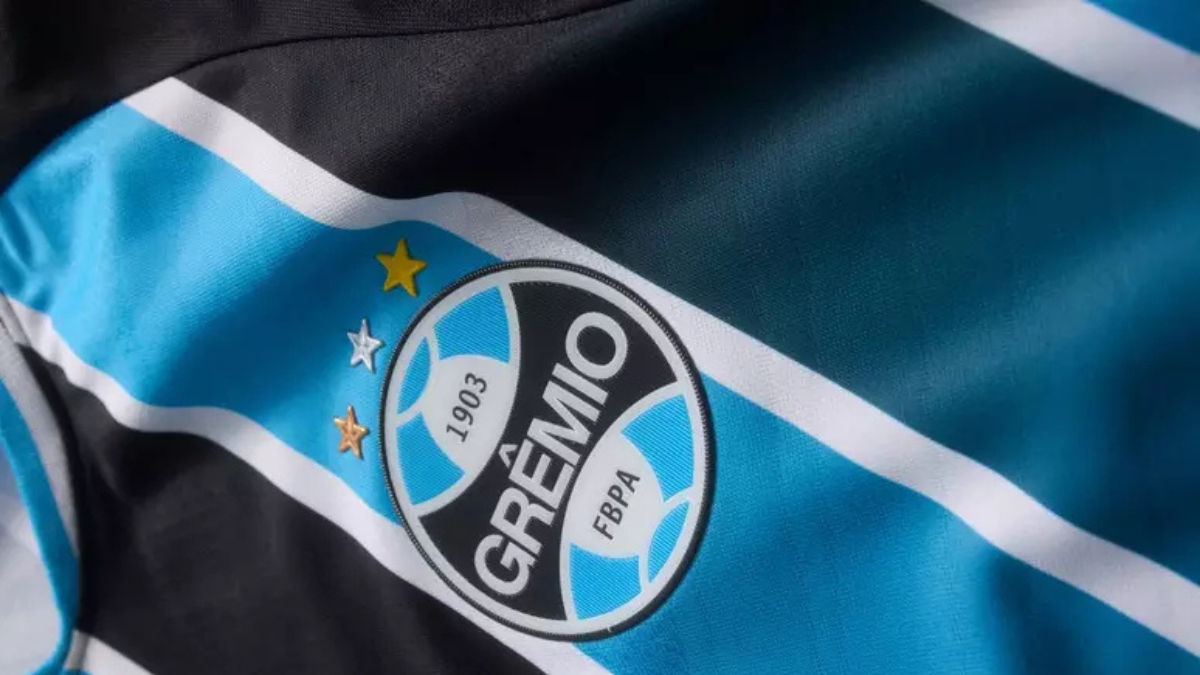 Bandeira do Grêmio tremulando ao vento, com listras azuis, pretas e brancas e o escudo do clube ao centro.