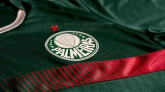Close-up do escudo do Palmeiras bordado em uma camisa de futebol verde escura, com uma estrela vermelha acima e um detalhe listrado em vermelho e preto na parte inferior.