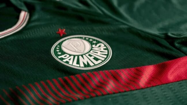 Close-up do escudo do Palmeiras bordado em uma camisa de futebol verde escura, com uma estrela vermelha acima e um detalhe listrado em vermelho e preto na parte inferior.