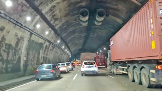 A imagem captura o intenso fluxo de veículos no interior de um túnel rodoviário, evidenciando o congestionamento típico de grandes deslocamentos. À direita, um grande caminhão transportando um contêiner vermelho domina parte da cena, enquanto diversas faixas de rolamento estão ocupadas por carros de passeio e outros veículos de carga que se estendem em direção ao fundo da estrutura. O túnel possui teto arqueado com sistemas de ventilação visíveis e iluminação artificial instalada nas laterais, refletindo sobre a pista e a carroceria dos automóveis. A composição transmite a sensação de lentidão e confinamento, característica de gargalos em rodovias importantes durante períodos de alta movimentação