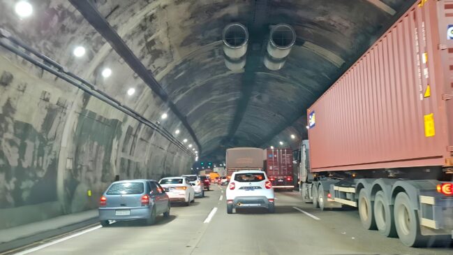 A imagem captura o intenso fluxo de veículos no interior de um túnel rodoviário, evidenciando o congestionamento típico de grandes deslocamentos. À direita, um grande caminhão transportando um contêiner vermelho domina parte da cena, enquanto diversas faixas de rolamento estão ocupadas por carros de passeio e outros veículos de carga que se estendem em direção ao fundo da estrutura. O túnel possui teto arqueado com sistemas de ventilação visíveis e iluminação artificial instalada nas laterais, refletindo sobre a pista e a carroceria dos automóveis. A composição transmite a sensação de lentidão e confinamento, característica de gargalos em rodovias importantes durante períodos de alta movimentação