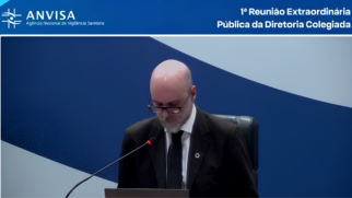 A imagem é uma captura de tela de uma transmissão oficial da Agência Nacional de Vigilância Sanitária (Anvisa). No centro da composição, um homem calvo, com barba grisalha e óculos de grau, aparece sentado e levemente inclinado para frente, mantendo o olhar baixo em direção a papéis ou a um notebook posicionado à sua frente. Ele veste um traje formal, composto por paletó preto, camisa branca e gravata escura. Além disso, um microfone de haste fina está direcionado para ele, indicando que o momento registra uma fala ou leitura oficial. O fundo é composto por um painel bicolor em tons de azul e branco, dividido por uma linha diagonal. Nesse sentido, na parte superior da imagem, há uma barra azul onde se lê, à esquerda, o logotipo da "ANVISA - Agência Nacional de Vigilância Sanitária" e, à direita, o texto identificador: "1ª Reunião Extraordinária Pública da Diretoria Colegiada". Desta maneira, a fotografia transmite a sobriedade e o caráter oficial do julgamento técnico realizado pelo órgão. Portanto, a imagem contextualiza o ambiente onde se decide o recurso contra a suspensão dos produtos da marca Ypê, evidenciando o processo institucional da agência.