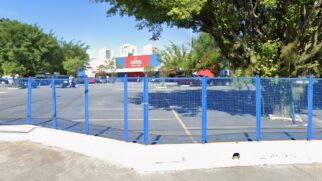 A imagem registra a fachada da unidade do supermercado Extra localizada na Avenida Senador Pinheiro Machado, em Santos. No registro, o logotipo vermelho e branco da rede se destaca sobre uma estrutura azul ao fundo, identificando o local onde funciona a Marapé Loterias, ponto da aposta premiada. Em primeiro plano, uma grade azul delimita o estacionamento, onde alguns veículos aparecem estacionados sob a luz de um dia ensolarado. A cena é composta por árvores frondosas e prédios residenciais ao fundo, elementos típicos do bairro Vila Mathias, o que confere autenticidade geográfica ao fato noticiado pelo Diário do Litoral.