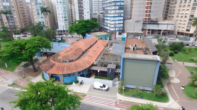 A imagem apresenta uma vista aérea do Aquário Municipal de Santos, localizado em uma área urbana densamente povoada. O edifício principal destaca-se por seu telhado de telhas cerâmicas alaranjadas com formato ovalado em uma das extremidades, contrastando com as paredes pintadas em um tom de azul vibrante. À direita da estrutura principal, observa-se um anexo de arquitetura mais moderna e retangular, também em azul, que possui uma grande fachada cega voltada para a rua.