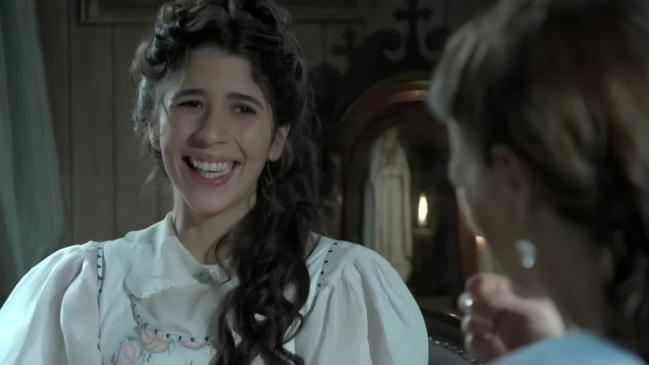 A imagem mostra a atriz Flora Diegues em uma cena da novela "Além do Tempo". Ela aparece em um plano médio, do peito para cima, caracterizada com um figurino de época do século XIX. A atriz está posicionada à esquerda, levemente voltada para a direita, exibindo um sorriso aberto e alegre enquanto olha para outra pessoa, que aparece apenas de costas e fora de foco no primeiro plano à direita. Flora tem cabelos castanhos escuros e ondulados, presos em um penteado elaborado com volume no topo. Ela veste uma blusa ou vestido branco com mangas bufantes e detalhes bordados em tons suaves de rosa e azul na parte frontal. O cenário ao fundo é escuro e rústico, sugerindo o interior de uma casa antiga com paredes de madeira. É possível notar um móvel de madeira entalhada e o que parece ser um espelho ou oratório ao fundo. A iluminação é suave e fria, destacando o rosto da atriz contra o ambiente sombrio