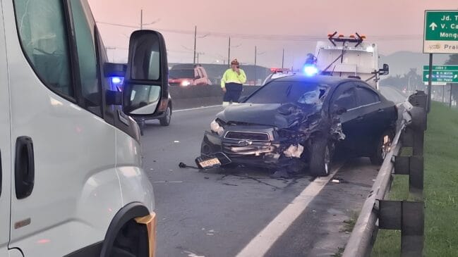 A imagem mostra as consequências de um grave acidente de trânsito em uma rodovia durante o amanhecer ou entardecer, dado o tom rosado do céu ao fundo. Em destaque, um automóvel de cor escura apresenta danos severos em toda a parte frontal esquerda, com a lataria completamente retorcida, o para-choque desprendido e o para-brisa estilhaçado pelo impacto. O veículo está parado próximo à mureta de proteção metálica da via, enquanto um funcionário da concessionária, vestindo uniforme fluorescente e capacete, sinaliza ou utiliza um rádio comunicador logo atrás do carro. É possível observar também um guincho com luzes de emergência ligadas posicionado para remover o veículo, além de placas de sinalização rodoviária indicando direções como Mongaguá e bairros de Praia Grande, o que reforça o contexto geográfico da ocorrência. No primeiro plano, à esquerda, a lateral de um veículo comercial branco compõe a cena, sugerindo que o tráfego no local está sendo monitorado ou desviado devido à gravidade da colisão.