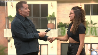 A fotografia registra um momento do reality show "A Casa do Patrão" durante uma transmissão ao vivo. Em primeiro plano, o apresentador Leandro Hassum aparece à esquerda, vestindo uma jaqueta azul-marinho de botões e usando um ponto eletrônico visível no ouvido. Ele segura um microfone com a mão direita enquanto aperta a mão da participante Nikita, que está posicionada à direita da imagem. Nikita sorri abertamente para Hassum, usando óculos de grau, um top preto estilo cropped e calças escuras com um cinto de fivela prateada. Nesse sentido, o cenário ao fundo simula a fachada de uma casa moderna com janelas grandes, sacadas de metal e vasos de plantas decorativos, iluminados por arandelas de luz quente. Além disso, no canto superior esquerdo da tela, lê-se a hashtag "#EliminaçãoNaCasa", enquanto no canto inferior direito aparece o selo informativo "AO VIVO". Dessa forma, a imagem captura o exato instante da despedida afetuosa entre o apresentador e a eliminada, momento que precedeu a intervenção do diretor Boninho pelos alto-falantes do estúdio. Portanto, a iluminação clara e a expressão descontraída dos dois personagens centrais contrastam com a tensão da bronca que ocorreria logo em seguida.