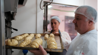 A imagem registra uma cena em plano médio dentro de uma cozinha industrial ou padaria. Dois profissionais, um homem e uma mulher, manipulam juntos uma assadeira repleta de massas de pães prontas para o cozimento. Elementos Principais (Primeiro Plano) O Padeiro: Posicionado no lado direito da imagem, um homem de pele clara, cabelos grisalhos nas laterais e perfil esquerdo visível segura a extremidade da assadeira. Ele está concentrado na atividade, usa uma touca descartável transparente na cabeça e veste um avental ou capa de proteção branca sobre a roupa. Suas mãos sustentam a base da bandeja metálica. A Assadeira e os Pães: Centralizada na metade inferior da imagem, uma grande assadeira retangular de metal envelhecido exibe diversas massas de pão cruas e amareladas. Na parte frontal, destacam-se três pães maiores modelados com cortes trançados na superfície. Atrás deles, há outras massas menores em formato de rolos cilíndricos alinhados. A assadeira está sendo direcionada para a abertura de um forno industrial de metal escuro, posicionado no canto esquerdo da cena. Elementos Secundários (Plano de Fundo) A Auxiliar ou Aluna: Logo atrás da assadeira, centralizada ao fundo, uma mulher de pele morena e cabelos presos sob uma touca descartável observa atentamente o movimento do colega. Ela veste uma blusa clara e um avental cinza com alças escuras, segurando levemente a borda oposta da bandeja para apoiar a manobra. O Ambiente: O cenário é composto pelas paredes brancas da cozinha, uma porta ou divisória de metal reflexivo ao fundo e a estrutura de aço do forno à esquerda. A iluminação é difusa e uniforme, focando a atenção na textura brilhante das massas cruas e no trabalho em equipe dos profissionais.
