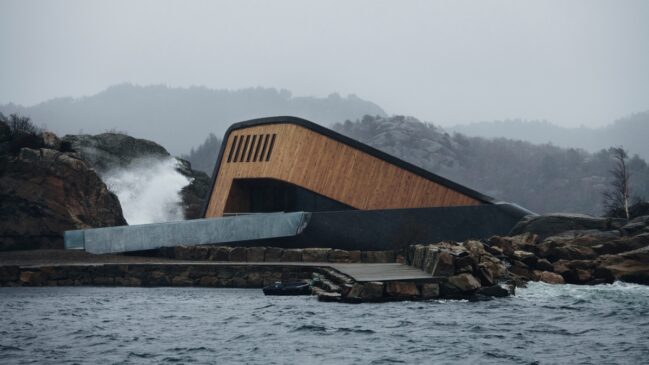 Edifício de arquitetura moderna e inclinada, com revestimento de madeira, construído sobre rochas na costa marítima.