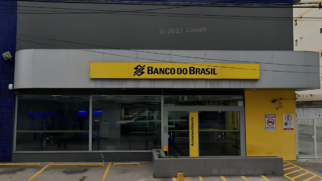 A imagem, capturada a partir do recurso Google Street View (com marca d'água "© 2025 Google"), mostra a fachada diurna de uma agência bancária do Banco do Brasil. A estrutura é moderna e apresenta as cores características da identidade visual da instituição financeira. Detalhes da Fachada: Letreiro Principal: No centro, fixado em uma estrutura de painel metálico cinza-claro, há um grande letreiro amarelo retangular com a inscrição "BANCO DO BRASIL" em letras escuras, precedido pelo logotipo da marca. Área de Autoatendimento: Abaixo do letreiro, grandes painéis de vidro transparente revelam o interior da agência. À esquerda, é possível visualizar uma fileira de caixas eletrônicos iluminados com luzes azuis. À direita, fica a porta de entrada de vidro com uma divisória amarela onde se lê verticalmente a palavra "Autoatendimento". Entorno e Sinalização: A parede da extrema esquerda é revestida por pequenas pastilhas azuis. A parede da extrema direita é pintada de amarelo e exibe placas de sinalização de trânsito/estacionamento (proibido estacionar motos e vaga regulamentada) ao lado do número do estabelecimento (870). Na frente da agência, há uma área de recuo pavimentada com marcações amarelas de vagas de estacionamento no chão, além de uma mureta baixa de blocos cinzas que funciona como floreira. Vários fios de energia cortam a parte superior da imagem, em frente à fachada cinza-escura do andar superior.