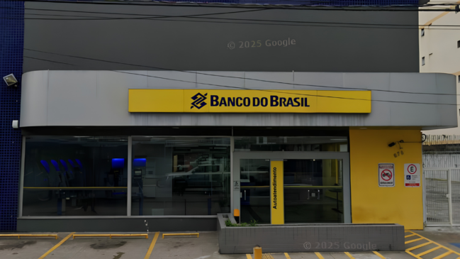 A imagem, capturada a partir do recurso Google Street View (com marca d'água "© 2025 Google"), mostra a fachada diurna de uma agência bancária do Banco do Brasil. A estrutura é moderna e apresenta as cores características da identidade visual da instituição financeira. Detalhes da Fachada: Letreiro Principal: No centro, fixado em uma estrutura de painel metálico cinza-claro, há um grande letreiro amarelo retangular com a inscrição "BANCO DO BRASIL" em letras escuras, precedido pelo logotipo da marca. Área de Autoatendimento: Abaixo do letreiro, grandes painéis de vidro transparente revelam o interior da agência. À esquerda, é possível visualizar uma fileira de caixas eletrônicos iluminados com luzes azuis. À direita, fica a porta de entrada de vidro com uma divisória amarela onde se lê verticalmente a palavra "Autoatendimento". Entorno e Sinalização: A parede da extrema esquerda é revestida por pequenas pastilhas azuis. A parede da extrema direita é pintada de amarelo e exibe placas de sinalização de trânsito/estacionamento (proibido estacionar motos e vaga regulamentada) ao lado do número do estabelecimento (870). Na frente da agência, há uma área de recuo pavimentada com marcações amarelas de vagas de estacionamento no chão, além de uma mureta baixa de blocos cinzas que funciona como floreira. Vários fios de energia cortam a parte superior da imagem, em frente à fachada cinza-escura do andar superior.