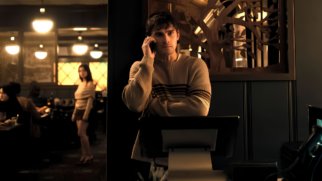 A imagem mostra uma captura de tela (still) de uma cena de filme em plano médio, ambientada no interior de um restaurante ou estabelecimento comercial com iluminação intimista e cinematográfica durante a noite. Elemento Principal (Primeiro Plano) O Homem ao Telefone: Um homem jovem, de pele clara e cabelos curtos e escuros, está posicionado do lado direito do centro da imagem. Ele está de pé, com o corpo ligeiramente voltado para a esquerda, e segura um smartphone preto junto ao ouvido direito, mantendo uma expressão séria e concentrada. Seus braços estão cruzados na altura do peito, apoiados sobre o que parece ser um balcão de atendimento ou monitor de caixa. Ele veste um suéter de tricô de gola careca na cor bege-claro, decorado com listras horizontais pretas e marrons na altura do tórax. O Cenário de Fundo Imediato: Atrás do homem, há um painel de parede escuro. À direita da sua cabeça, destaca-se um elemento decorativo vazado de madeira escura com o formato de galhos de uma árvore estilizada. Segundo Plano (Fundo à Esquerda) A Mulher Observando: Através de uma abertura ou passagem à esquerda do homem, avista-se um segundo ambiente do restaurante. Uma mulher jovem, de pele clara e longos cabelos escuros, está de pé e ligeiramente desfocada. Ela veste uma blusa bege de ombros caídos e uma saia curta escura. O seu olhar está direcionado de perfil em direção ao homem do primeiro plano. O Salão do Restaurante: Mais ao fundo e à esquerda, observam-se silhuetas de clientes sentados em mesas sob uma iluminação amarelada e aconchegante. Luminárias esféricas pendentes de estilo retrô estão acesas no teto, criando pontos de luz circular contra as janelas quadriculadas do fundo. Iluminação e Cores Paleta de Cores: A imagem apresenta uma atmosfera dramática e aconchegante, dominada por tons terrosos, pretos, cinzas e marrons, contrastados pelo brilho dourado e quente das lâmpadas. Efeito de Luz: A iluminação foca predominantemente no rosto e nos ombros do homem em primeiro plano, deixando as áreas laterais e os cantos inferiores em sombras profundas (penumbra).