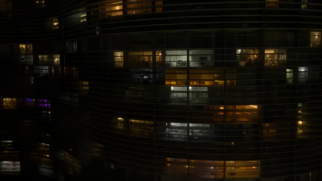 A imagem mostra a fachada externa de um grande edifício residencial iluminado à noite, capturada a partir de um ângulo frontal e ligeiramente elevado. O prédio possui linhas horizontais contínuas e onduladas formadas por brises ou vigas de concreto, que cruzam toda a extensão da imagem, criando um padrão geométrico e sinuoso característico da arquitetura moderna. A estrutura do edifício faz uma leve curva da esquerda para a direita, sugerindo um formato em "S". A maior parte da fachada está na penumbra, mas dezenas de janelas retangulares de apartamentos estão iluminadas por dentro. As luzes internas variam de intensidade e tom: algumas janelas emitem um brilho amarelado e quente, enquanto outras mostram uma luz branca e fria. É possível notar silhuetas vagas de móveis, divisórias e tetos através dos vidros de alguns apartamentos, revelando fragmentos da vida urbana e doméstica dos moradores no interior da estrutura vertical.