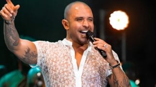 A imagem retrata o cantor Diogo Nogueira durante uma performance ao vivo. Ele aparece sorridente e com uma expressão alegre, segurando um microfone preto com a mão esquerda enquanto levanta o braço direito, apontando para cima com o dedo indicador. O artista veste uma camisa branca de renda transparente com bordados florais delicados, revelando suas diversas tatuagens nos braços. Ele também usa acessórios como um anel de destaque, uma pulseira metálica e um relógio. A iluminação de palco cria um efeito de brilho em seu rosto e destaca o fundo escuro, onde se percebe a presença de outros músicos e equipamentos de iluminação em tons esverdeados, mantendo o foco central na figura do sambista.