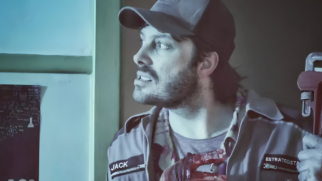A imagem apresenta o humorista e apresentador Danilo Gentili em uma cena do filme "Os Exterminadores do Além contra a Loira do Banheiro". O ator aparece de perfil com uma expressão de tensão e utiliza um boné escuro junto a um uniforme cinza de trabalho. No traje, é possível identificar as identificações "JACK" e "ESTRATEGISTA" bordadas no peito. Ele segura uma chave de grifo vermelha e exibe as mãos manchadas com sangue artificial, o que reforça o gênero de comédia de horror da obra. A composição conta com uma iluminação fria e azulada que destaca o protagonista contra o fundo escuro do cenário.