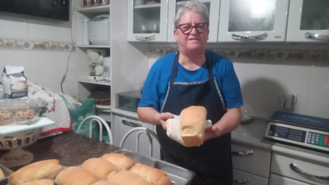 A imagem captura uma senhora de óculos e cabelos grisalhos em uma cozinha, segurando com orgulho um pão caseiro recém-assado. Ela veste uma camiseta azul sob um avental escuro, posicionando-se à frente de uma bancada onde repousa uma assadeira repleta de outros pães dourados e fofos. O cenário ao fundo revela uma cozinha doméstica bem equipada, com armários brancos, prateleiras organizadas com eletrodomésticos como batedeira e cafeteira, além de uma balança digital à direita, sugerindo um ambiente de produção cuidadosa e artesanal. A iluminação clara destaca a textura dos pães e o semblante satisfeito da mulher, transmitindo uma sensação acolhedora de culinária feita com dedicação.