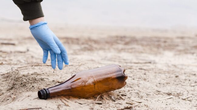 Garrafa de plástico jogada na faixa de areia sendo recolhida