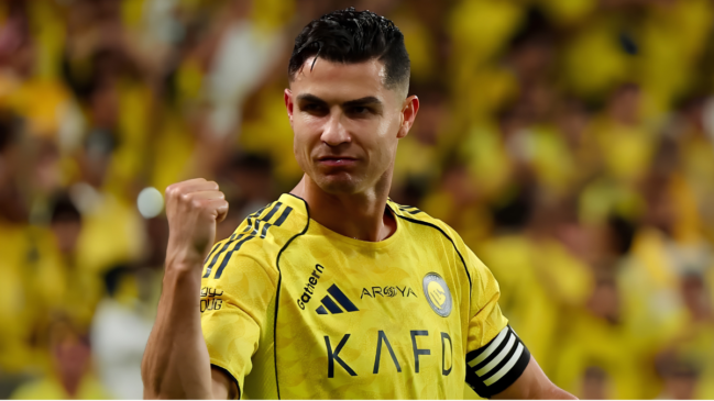 Esta imagem apresenta um retrato em plano médio e focado no jogador de futebol Cristiano Ronaldo, capturado em um momento de celebração ou determinação dentro do campo. O Personagem e a Expressão O atleta aparece centralizado e ligeiramente voltado para a esquerda da imagem. Ele exibe uma expressão facial intensa e compenetrada, com os olhos semi-cerrados fixos à frente, lábios pressionados e testa franzida, transmitindo uma postura de foco e autoconfiança. O jogador ergue o braço direito com o punho cerrado na altura do peito, em um gesto clássico de comemoração ou vibração. O cabelo está curto, cortado em estilo degradê nas laterais e penteado com gel para trás na parte superior. Vestuário e Detalhes Ele veste a camisa oficial amarela do clube saudita Al-Nassr, detalhada com linhas pretas finas que demarcam os ombros e a gola redonda. No peito, destacam-se o logotipo preto da fornecedora esportiva Adidas, a inscrição do patrocinador secundário "Aroya" e o brasão redondo do clube em tons de amarelo e azul no lado esquerdo. A estampa principal exibe as letras estilizadas "KAFD" em preto na região central. No braço esquerdo, nota-se o detalhe da braçadeira de capitão com listras pretas e brancas. O Segundo Plano O fundo da imagem mostra as arquibancadas do estádio completamente lotadas. A torcida surge de forma desfocada (efeito bokeh), criando uma grande massa de tons amarelos vibrantes que destaca a silhueta do jogador em primeiro plano.