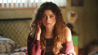 A imagem destaca a atriz Zendaya interpretando sua personagem Rue Bennett na série Euphoria da HBO. Ela aparece sentada em um quarto, com uma expressão pensativa e o olhar direcionado para cima. A personagem possui cabelos longos, escuros e cacheados que caem sobre os ombros, e veste um casaco de moletom vinho sobre uma regata escura. O cenário ao fundo apresenta uma iluminação suave e amarelada, revelando detalhes de um quarto adolescente com elementos desfocados, como a cabeceira da cama e objetos em uma cômoda. A composição da cena foca no rosto da atriz, transmitindo o tom introspectivo e dramático característico da produção.