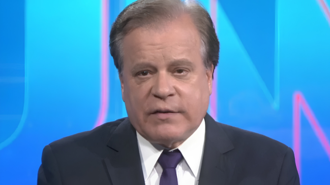 A imagem apresenta um plano médio do jornalista Chico Pinheiro, conhecido por sua longa trajetória na Rede Globo. Ele aparece vestido formalmente com um paletó azul-marinho, camisa social branca e uma gravata roxa. O jornalista mantém uma expressão séria e concentrada, característica de sua atuação em telejornais. Ao fundo, observa-se um cenário de estúdio televisivo em tons de azul brilhante, com elementos gráficos desfocados que remetem à identidade visual de programas de notícias. A iluminação é frontal e nítida, destacando os traços do apresentador e reforçando o tom profissional e informativo da cena.