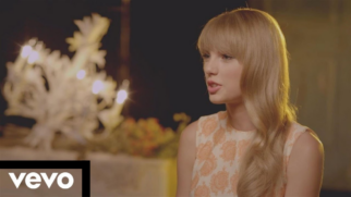 A imagem mostra um plano médio da cantora Taylor Swift em um cenário iluminado de forma suave, com a marca d'água da Vevo no canto inferior esquerdo. Taylor Swift A cantora está posicionada do centro para o lado direito da tela, de perfil em três quartos, voltada para a esquerda. Ela tem pele clara e cabelos loiros longos, ondulados, com uma franja reta que cobre a testa. Sua boca está levemente aberta, como se estivesse falando. Ela veste um vestido sem mangas de tom claro, estampado com padrões florais ou arabescos em relevo na cor laranja claro. Cenário e Iluminação O fundo está obscurecido e fora de foco (efeito bokeh). Do lado esquerdo, atrás da cantora, há uma luminária ou candelabro decorativo com lâmpadas pequenas acesas que emitem um brilho suave e quente. Abaixo das luzes, notam-se algumas flores ou arranjos desfocados. A iluminação geral é dourada e incide suavemente sobre o rosto e o cabelo da artista. Elementos Gráficos No canto inferior esquerdo da imagem, há um retângulo preto horizontal que serve de base para o logotipo oficial da "vevo", escrito em letras minúsculas na cor branca.