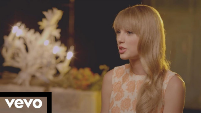 A imagem mostra um plano médio da cantora Taylor Swift em um cenário iluminado de forma suave, com a marca d'água da Vevo no canto inferior esquerdo. Taylor Swift A cantora está posicionada do centro para o lado direito da tela, de perfil em três quartos, voltada para a esquerda. Ela tem pele clara e cabelos loiros longos, ondulados, com uma franja reta que cobre a testa. Sua boca está levemente aberta, como se estivesse falando. Ela veste um vestido sem mangas de tom claro, estampado com padrões florais ou arabescos em relevo na cor laranja claro. Cenário e Iluminação O fundo está obscurecido e fora de foco (efeito bokeh). Do lado esquerdo, atrás da cantora, há uma luminária ou candelabro decorativo com lâmpadas pequenas acesas que emitem um brilho suave e quente. Abaixo das luzes, notam-se algumas flores ou arranjos desfocados. A iluminação geral é dourada e incide suavemente sobre o rosto e o cabelo da artista. Elementos Gráficos No canto inferior esquerdo da imagem, há um retângulo preto horizontal que serve de base para o logotipo oficial da "vevo", escrito em letras minúsculas na cor branca.