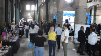 Uma imagem colorida, em plano médio e em nível dos olhos, mostra o interior de um grande armazém reformado com um piso de concreto polido e uma mistura de pilares de aço preto e arcos de pedra de calcário cinza, com o teto em altura tripla e sem acabamento. Duas fileiras de pilares de metal preto e verde escuro estendem-se até o fundo do salão, em direção a um arco de pedra que revela uma saída para um pátio ensolarado. No centro do corredor, estão dispostas fileiras de mesas de exibição que vendem artesanato, artesanato em joias, tecidos e roupas. Uma multidão de cerca de 30 a 40 pessoas está misturada e caminha pelo local, visualizando as exposições e as demonstrações nas mesas. Três mulheres e dois homens em primeiro plano, todos de costas para nós, caminham para longe do corredor central, visualizando as exposições. À direita, um pequeno palco e equipamento de som foram montados, juntamente com um arco de balões azuis, no qual cinco homens vestidos de preto e cinza ficam de frente um para o outro e conversam. A luz do dia flui através de duas grandes aberturas de janela arqueadas na parede oposta e de um pátio ao ar livre no fundo, com as janelas e o pátio ao fundo aparecendo muito brilhantes devido à luz solar natural e os elementos do teto superior aparecendo como formas e contornos suaves e escuras.