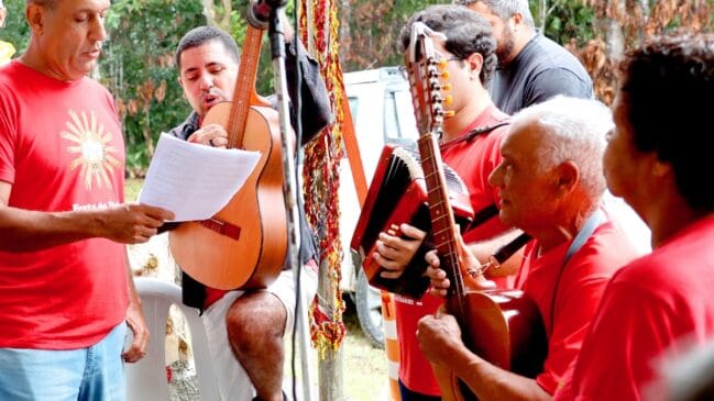 Fotografia colorida de um grupo de homens cantando e tocando instrumentos musicais ao ar livre. Eles usam camisetas vermelhas com o símbolo do Divino Espírito Santo. Um homem à esquerda lê a letra em um papel, enquanto os outros tocam violão, sanfona e viola