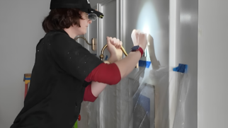 A imagem mostra uma fotografia em plano médio de perfil esquerdo, registrando um jovem profissional trabalhando na instalação ou ajuste de uma luminária de parede em um ambiente interno residencial durante o dia. Elemento Principal (Primeiro Plano) O Profissional em Atividade: Um jovem de pele clara e cabelos castanhos na altura do pescoço está posicionado do lado esquerdo, voltado para a direita. Ele está concentrado e com os dois braços erguidos na altura do peito, manipulando uma peça metálica fixada na parede. Vestimentas e Acessórios: Ele usa um boné preto com uma lanterna de cabeça acoplada na aba, apontando um feixe de luz brilhante diretamente para a área de trabalho na parede. Veste uma camisa preta de mangas curtas por cima de uma blusa de mangas compridas na cor vermelha, cujas mangas estão puxadas até os antebraços. No pulso esquerdo, destaca-se um relógio inteligente (smartwatch) com pulseira vermelha. Cenário e Detalhes Técnicos (Plano de Fundo) A Parede e a Luminária: A parede ao fundo é branca e possui painéis decorativos com molduras em relevo (estilo boiserie). O jovem segura um braço de metal tubular curvado e dourado, que faz parte de uma arandela de iluminação. Uma estrutura semelhante já instalada pode ser vista logo atrás de seu ombro. Proteção do Ambiente: Abaixo da luminária em manutenção, a parede está coberta por uma lona plástica protetora transparente, fixada horizontalmente com pedaços de fita adesiva de pintor na cor azul, protegendo a superfície contra poeira ou arranhões. No canto inferior esquerdo, vê-se a parte superior de uma escada doméstica de metal com detalhes em laranja e azul. Iluminação da Cena: A iluminação geral do ambiente é clara, mas o grande destaque visual é o círculo de luz focado e intenso gerado pela lanterna de cabeça do profissional, que clareia fortemente a parede branca à sua frente.