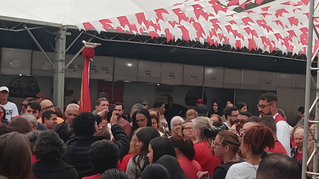 Em uma tomada fotográfica em plano médio e horizontal, tirada de trás para frente, uma multidão se aglomera sob uma grande tenda branca de lona durante um evento que aparenta ser uma celebração religiosa ou cultural popular. Detalhes da Multidão e Vestimentas Composição: O público é diversificado, composto por homens e mulheres de várias idades, predominantemente adultos e idosos. A maioria está de costas ou de perfil para a câmera, olhando em direção ao centro e ao lado direito da imagem. Cores: Muitas pessoas na multidão vestem camisetas na cor vermelha, criando blocos de cor marcantes em meio a outras roupas brancas, pretas e cinzas. Ações e Expressões: No lado esquerdo, um homem de costas levanta um celular com a mão direita para registrar o momento. Mais à direita, um jovem de óculos e cabelos curtos, vestindo uma túnica branca com uma estola vermelha sobre os ombros — vestimenta típica de seminarista, coroinha ou clérigo —, observa a movimentação. Próximo a ele, um homem segura uma câmera de vídeo profissional apoiada no ombro, sugerindo a cobertura da imprensa ou registro oficial do evento. Elementos Culturais e Religiosos A Bandeira do Divino: À esquerda do centro, ergue-se um longo mastro vertical com uma bandeira ou estandarte de tecido vermelho e detalhes dourados. No topo do mastro, acima de um arranjo de tecido vermelho em formato esférico, destaca-se a escultura de uma pequena pomba branca de asas abertas, símbolo tradicional do Divino Espírito Santo. Decoração: O teto da grande tenda está ricamente decorado com dezenas de varais paralelos repletos de bandeirinhas de festa de papel ou plástico, alternadas rigidamente nas cores branca e vermelha. O Cenário ao Fundo Ao fundo da multidão, sob a cobertura da tenda, alinham-se divisórias de stands ou estandes comerciais temporários na cor cinza-clara. Em um dos stands do lado esquerdo, é possível ler parcialmente um letreiro em letras escuras com a inscrição "Auto Peças", indicando que a celebração acontece em um espaço de feira, praça pública ou pavilhão comunitário. No canto inferior direito, a estrutura metálica de treliça de uma torre de sustentação (box truss) delimita o enquadramento. A iluminação é difusa e natural, típica de um dia nublado sob cobertura de lona.