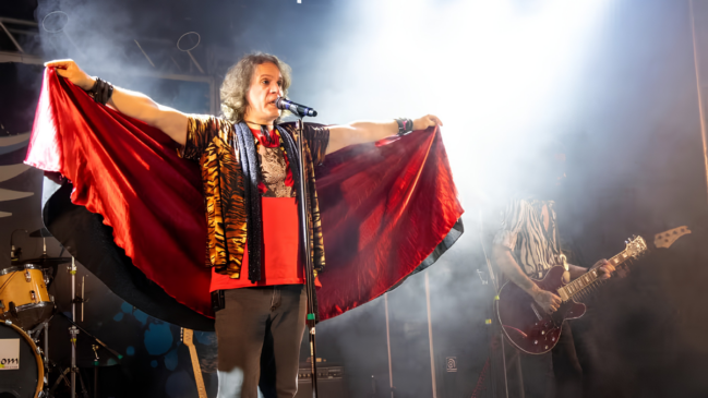 Uma fotografia de plano médio e horizontal captura uma apresentação de rock ao vivo em um palco coberto por fumaça cenográfica e iluminação artística. Elementos em Destaque O Cantor (Centro do Palco): Um homem de pele clara e cabelos castanhos ondulados, na altura dos ombros, canta em um microfone fixado em um pedestal. Ele está com os braços totalmente abertos para os lados, segurando e estendendo uma longa capa de cetim vermelho-escuro com o forro preto. O vocalista veste uma camiseta vermelha sob um colete com estampa de tigre (listras pretas e amarelas) e calça cinza-escura. Ele usa munhequeiras pretas em ambos os pulsos e um colar preto justo ao pescoço. O Guitarrista (Lado Direito): Em segundo plano e parcialmente envolto por fumaça, um integrante da banda toca uma guitarra elétrica de corpo semiacústico na cor vinho (vermelho-escura). Ele tem barba, veste uma camisa de manga curta com estampa listrada em preto e branco (estilo zebra) e calça escura. A Bateria (Canto Inferior Esquerdo): Uma parte do kit de bateria é visível, mostrando um tom-tom de tom amarelo-claro e a lateral do bumbo com uma pele branca, além dos pedestais de metal dos pratos. Iluminação, Cores e Clima Efeitos e Luz: Um forte feixe de luz branca vem de trás e do alto (contra-luz) no lado direito, criando um efeito de silhueta e iluminando intensamente a fumaça branca que preenche o ambiente do palco. Cores: O vermelho vivo da capa e da camiseta do cantor cria o principal ponto de contraste visual contra o fundo escuro e a névoa cinzenta do palco. Ambiente: A atmosfera evoca a energia e a dramaticidade de um show de rock clássico ou performance teatral tributo.