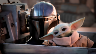 A imagem apresenta um plano médio em formato paisagem com foco centralizado nos dois personagens principais da franquia Star Wars, o Mandaloriano Din Djarin e a criatura Grogu, acomodados dentro de uma estrutura metálica que remete ao cockpit de uma espaçonave ou veículo de transporte. Elementos Principais (Primeiro Plano) O Mandaloriano (Din Djarin): Posicionado ligeiramente à esquerda do centro, o guerreiro veste seu icônico capacete metálico prateado e brilhante feito de beskar. O metal reflete nuances sutis de luz quente vindas do ambiente externo. A viseira em formato de "T" é completamente preta e opaca, ocultando seu rosto. Ele está voltado para a frente, em uma postura firme e protetora ao lado de seu companheiro. Grogu (Baby Yoda): Posicionado à direita, em primeiro plano, a pequena criatura alienígena aparece de corpo inteiro, levemente inclinada para a esquerda. Ele tem a pele verde-clara, olhos negros grandes e expressivos, e orelhas longas e pontiagudas estendidas para as laterais. Veste sua tradicional túnica de tecido grosso na cor bege com gola alta e felpuda. Sua mãozinha esquerda está estendida para a frente com os pequenos dedos abertos, enquanto ele olha sutilmente em direção ao espectador. Cenário e Detalhes Técnicos (Plano de Fundo) O Veículo: Os personagens estão abrigados atrás de uma placa horizontal de metal escovado e fosco, que corta a parte inferior da imagem. À esquerda de Din Djarin, ergue-se parte da estrutura interna do veículo, composta por peças mecânicas, tubulações e painéis em tons de cinza e bege industrial. O Fundo e Iluminação: O plano de fundo está fortemente desfocado (efeito bokeh), revelando um ambiente externo árido ou urbano em tons de marrom e areia, sugerindo um planeta desértico. A iluminação é suave e cinematográfica, concentrando os pontos de brilho no topo do capacete do Mandaloriano e nos contornos do rosto e das orelhas de Grogu.