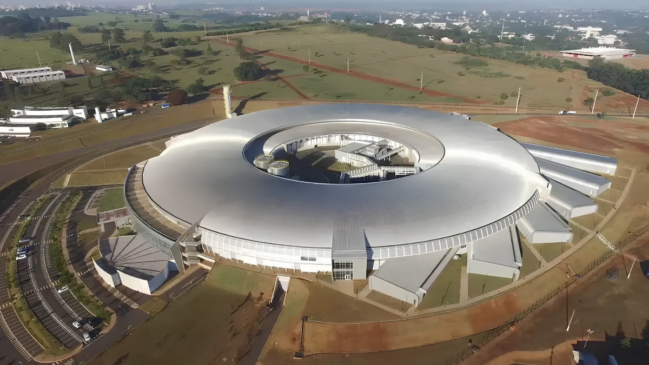 O Prédio Principal (Sirius) O grande destaque da foto é a gigantesca estrutura arquitetônica de formato circular e futurista, que lembra um grande anel ou um disco voador pousado no solo. O edifício possui uma cobertura metálica em tom cinza-prateado claro, dividida em gomos concêntricos que convergem para um pátio central descoberto. Na fachada frontal, há uma entrada principal envidraçada de pé-direito alto, conectada a pequenas estruturas anexas e passarelas que se projetam para fora do anel principal. Ao lado esquerdo da construção, estende-se um grande estacionamento pavimentado em formato semicircular, com diversas vagas marcadas e alguns carros visíveis. O Entorno e a Paisagem O complexo laboratorial está inserido em um terreno amplo, cercado por vastas áreas de gramado e terra avermelhada, com algumas árvores esparsas e vias internas de asfalto que contornam os prédios. No quadrante superior esquerdo, avistam-se outras instalações industriais e laboratoriais menores de cor branca. Ao fundo da composição, a paisagem se divide entre campos abertos de vegetação rasteira e, mais ao longe, a linha do horizonte revela a malha urbana de uma cidade, com prédios e casas sob um céu claro e ensolarado.