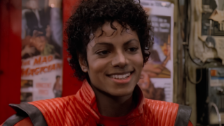 A imagem apresenta um close focado em Michael Jackson, capturado durante a era do icônico videoclipe "Thriller". O cantor aparece sorrindo, exibindo seus dentes e um olhar brilhante, enquanto ostenta o penteado de cachos curtos e úmidos, conhecido como estilo jheri curl, que foi sua marca registrada no início dos anos 80. Ele utiliza a famosa jaqueta de couro vermelha com detalhes e gola pretos, peça que se tornou um símbolo cultural global. O cenário ao fundo, embora levemente desfocado, revela o interior de uma sala ou corredor com cartazes de filmes antigos fixados em uma parede vermelha, sendo possível identificar parcialmente o título "The Mad Magician" em um dos pôsteres. Nesta versão, a figura feminina que estava em primeiro plano à direita na fotografia original foi removida, deixando o enquadramento totalmente centralizado no artista e preservando a iluminação quente e a estética cinematográfica da produção de 1983.