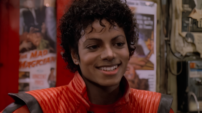 A imagem apresenta um close focado em Michael Jackson, capturado durante a era do icônico videoclipe "Thriller". O cantor aparece sorrindo, exibindo seus dentes e um olhar brilhante, enquanto ostenta o penteado de cachos curtos e úmidos, conhecido como estilo jheri curl, que foi sua marca registrada no início dos anos 80. Ele utiliza a famosa jaqueta de couro vermelha com detalhes e gola pretos, peça que se tornou um símbolo cultural global. O cenário ao fundo, embora levemente desfocado, revela o interior de uma sala ou corredor com cartazes de filmes antigos fixados em uma parede vermelha, sendo possível identificar parcialmente o título "The Mad Magician" em um dos pôsteres. Nesta versão, a figura feminina que estava em primeiro plano à direita na fotografia original foi removida, deixando o enquadramento totalmente centralizado no artista e preservando a iluminação quente e a estética cinematográfica da produção de 1983.