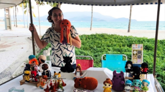 Uma fotografia colorida, de plano médio e em nível dos olhos, mostra uma mulher branca, de cabelos escuros presos e usando óculos de grau, sorrindo e segurando um brinquedo de polvo de crochê laranja e amarelo. Ela veste uma camiseta branca com estampa abstrata em azul e verde. A mulher está atrás de uma mesa coberta com um pano branco e repleta de diversos brinquedos de crochê artesanais, organizados em fileiras. Os brinquedos retratam personagens famosos da cultura pop, incluindo super-heróis como Wolverine, Deadpool e Pantera Negra; personagens de animação como Goku (Dragon Ball), Pikachu (Pokémon), Bela (A Bela e a Fera) e Totoro (Meu Amigo Totoro); e outros bonecos originais. Há também pequenas placas com informações de preço e códigos QR ao lado dos bonecos, e uma garrafa de água atrás. A cena se passa em uma barraca de feira em uma praia, com a areia clara e o mar azul claro ao fundo. Algumas palmeiras e montanhas ao longe completam a paisagem. No canto superior esquerdo, há uma lona de teto de cor bege com detalhes em azul escuro e um mastro. O chão é de cimento com textura de pedras pequenas.