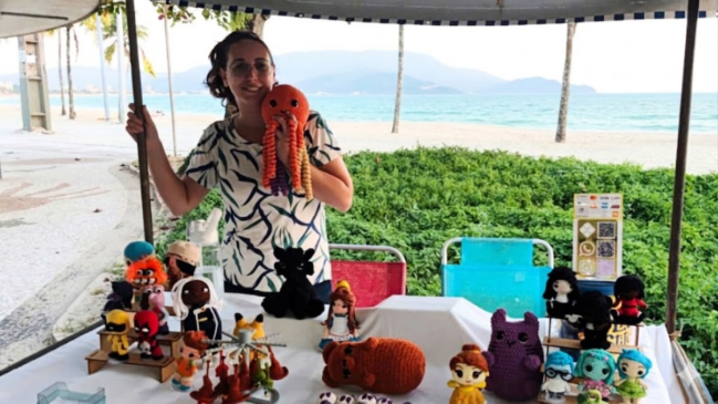 Uma fotografia colorida, de plano médio e em nível dos olhos, mostra uma mulher branca, de cabelos escuros presos e usando óculos de grau, sorrindo e segurando um brinquedo de polvo de crochê laranja e amarelo. Ela veste uma camiseta branca com estampa abstrata em azul e verde. A mulher está atrás de uma mesa coberta com um pano branco e repleta de diversos brinquedos de crochê artesanais, organizados em fileiras. Os brinquedos retratam personagens famosos da cultura pop, incluindo super-heróis como Wolverine, Deadpool e Pantera Negra; personagens de animação como Goku (Dragon Ball), Pikachu (Pokémon), Bela (A Bela e a Fera) e Totoro (Meu Amigo Totoro); e outros bonecos originais. Há também pequenas placas com informações de preço e códigos QR ao lado dos bonecos, e uma garrafa de água atrás. A cena se passa em uma barraca de feira em uma praia, com a areia clara e o mar azul claro ao fundo. Algumas palmeiras e montanhas ao longe completam a paisagem. No canto superior esquerdo, há uma lona de teto de cor bege com detalhes em azul escuro e um mastro. O chão é de cimento com textura de pedras pequenas.