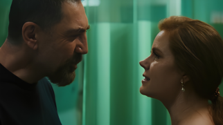 A imagem registra uma cena dramática em primeiro plano, destacando o confronto direto e tenso entre um homem e uma mulher dispostos de perfil, um de frente para o outro. Elementos Principais (Primeiro Plano) O Homem (Lado Esquerdo): O ator Javier Bardem aparece posicionado de perfil, voltado para o lado direito. Ele tem cabelos curtos e escuros, além de uma barba rala com cavanhaque bem marcado. Sua expressão facial é agressiva e intimidadora, com os olhos semicerrados e a testa franzida, demonstrandohostilidade física próxima em direção à mulher. Ele veste uma camiseta preta simples. A Mulher (Lado Direito): A atriz Amy Adams está posicionada também de perfil, voltada para o lado esquerdo, encarando o homem. Ela tem cabelos ruivos amarrados em um rabo de cavalo baixo e usa um brinco dourado delicado. Sua expressão mistura tensão e firmeza, com os olhos bem abertos e a boca levemente entreaberta, sugerindo uma reação ou enfrentamento verbal em meio ao confronto. Elementos Secundários (Plano de Fundo) O Cenário: O fundo da cena é composto por uma divisória ou cortina hospitalar translúcida em um tom verde-esmeralda bem característico, disposta em faixas verticais. A Iluminação: A iluminação é difusa e concentrada nos rostos dos personagens, criando sombras marcadas que acentuam o clima de suspense e a gravidade do momento dramático capturado.