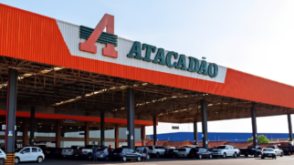 A imagem mostra uma fotografia em plano médio e ângulo frontal da fachada externa de uma grande loja de hipermercado da rede Atacadão, registrada durante um dia claro com céu azul aberto. Estrutura da Fachada A Platibanda Superior (Cobertura): O topo do edifício é composto por uma longa e alta estrutura metálica horizontal. As extremidades laterais e as bordas dessa cobertura são pintadas em um tom de laranja vivo. O Painel Central da Marca: No centro dessa platibanda, há um painel de fundo metálico branco frisado. Sobre ele, está fixado o logotipo oficial da empresa. À esquerda, destaca-se a letra estilizada "A" em tamanho grande e cor laranja, cortada por três listras horizontais verdes. À direita do logotipo, está escrita a palavra "ATACADÃO" em letras maiúscicas e na cor verde-escuro. Área Térrea e Estacionamento O Pátio Coberto (Marquise): Abaixo da grande estrutura laranja, estende-se uma ampla cobertura suspensa suportada por robustos pilares verticais de metal escuro. Essa marquise projeta uma sombra sobre a área de circulação e as vagas de estacionamento frontais. O teto interno da estrutura revela vigas metálicas entrelaçadas. Os Veículos: Diversos carros de passeio de diferentes modelos e cores (incluindo branco, prata, cinza e preto) estão estacionados em fila indiana logo abaixo da cobertura. No canto esquerdo, um carro compacto branco ganha destaque. No canto direito, caminhonetes e um caminhão pequeno estão posicionados na área descoberta. A Circulação: É possível observar poucas silhuetas de pessoas caminhando entre os veículos no pátio. Ao fundo, após o limite do estacionamento, nota-se uma parede azul de outro galpão industrial e o contorno de algumas árvores sob o céu limpo.