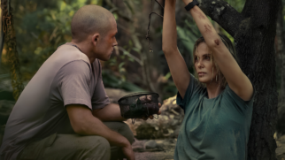 Esta imagem apresenta uma cena dramática em meio a um ambiente de floresta densa e sombreada, com foco em dois personagens. À direita, uma mulher loira e com cabelos molhados está sentada no chão, encostada no tronco de uma árvore. Ela veste uma camiseta verde-escura suja de poeira e tem os braços erguidos e amarrados por uma corda a um galho acima. Há pequenos ferimentos e marcas de sangue em seus braços e bochecha, e sua expressão é de exaustão e seriedade. À esquerda, um homem com a cabeça raspada e vestindo camiseta e calça camuflada de tom cáqui está agachado de perfil, olhando fixamente para a mulher. Ele segura uma pequena cesta de metal preta com algumas plantas e o que parecem ser pequenos frutos ou mantimentos em seu interior. O fundo mostra uma vegetação rústica e desfocada, sugerindo um cenário de sobrevivência ou cativeiro na mata.