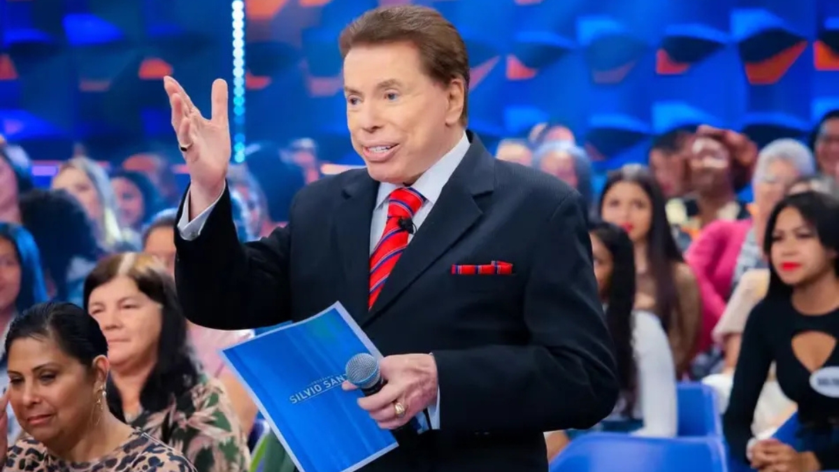 Fotografia do apresentador Silvio Santos no palco de seu programa vestindo terno preto e gravata listrada vermelha gesticulando com a mão direita erguida enquanto segura um microfone e um cartão azul