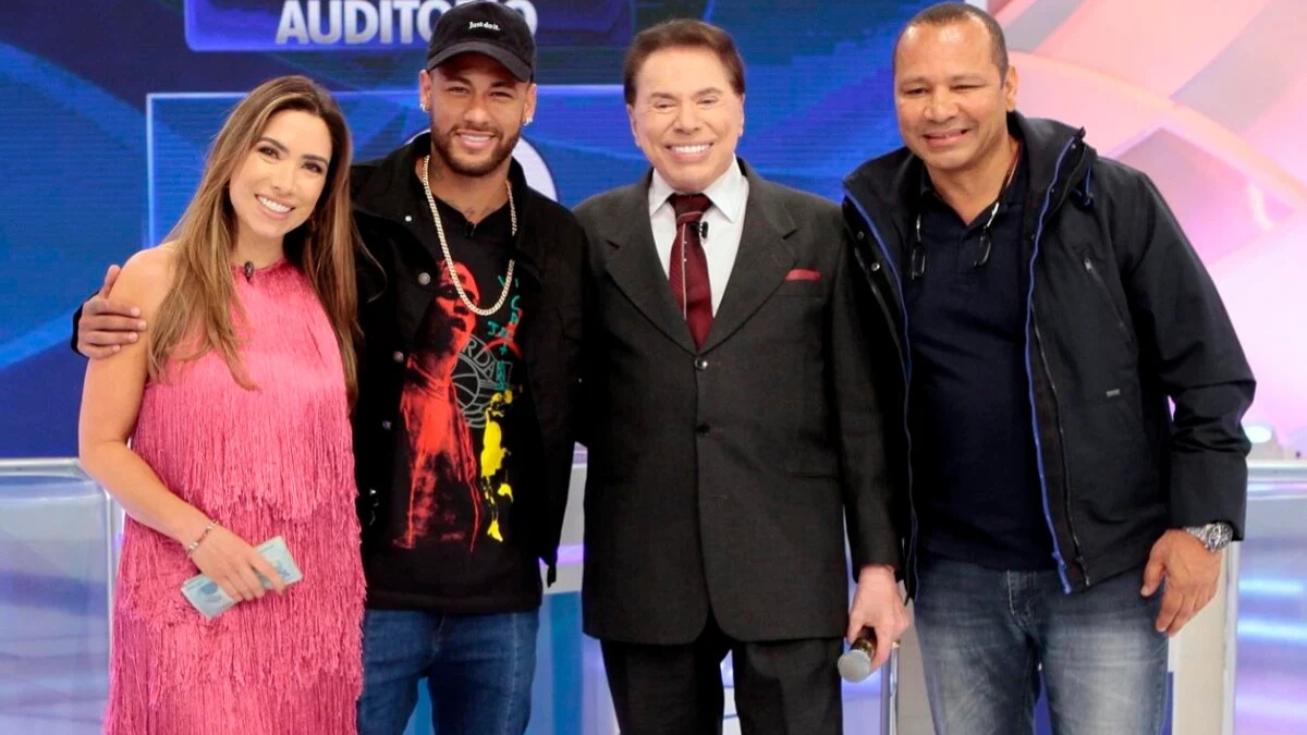 Fotografia em plano médio mostrando Patrícia Abravanel, o jogador Neymar Jr, o apresentador Silvio Santos e Neymar Pai sorrindo juntos no palco de um programa de televisão