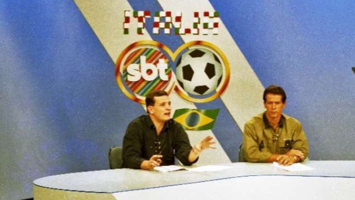 Fotografia de arquivo de dois apresentadores sentados na bancada do estúdio do SBT durante a cobertura da Copa do Mundo da Itália, com o logotipo da emissora e uma bola de futebol ao fundo
